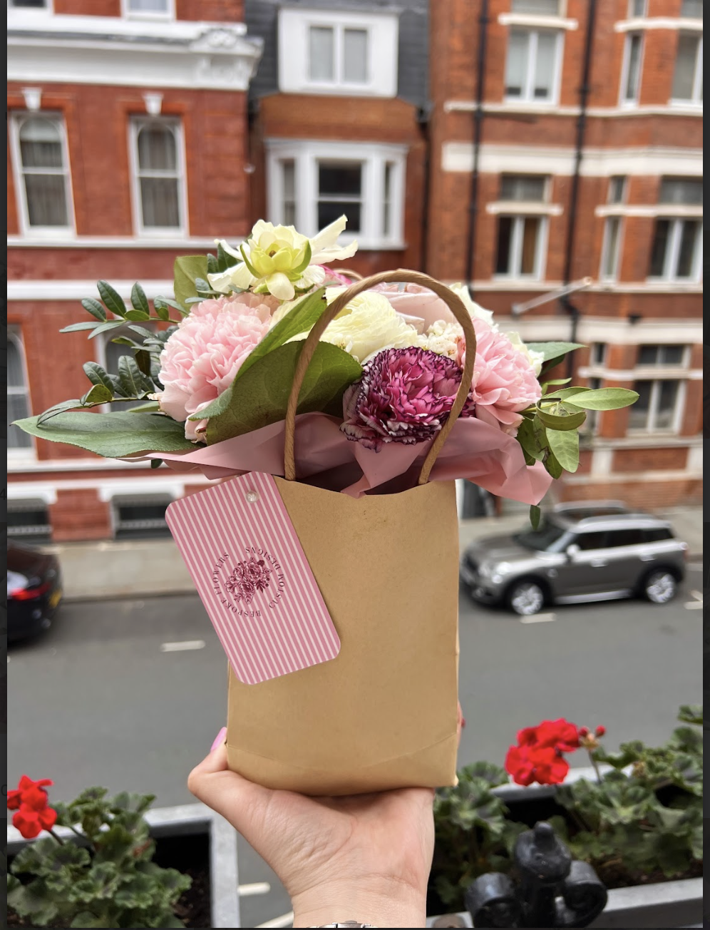 A person's hand holding a small paper gift bag decorated with pink and white striped tag, containing a bouquet of pink, white, and purple flowers with green leaves, against a backdrop of a city street with red brick buildings and parked cars.