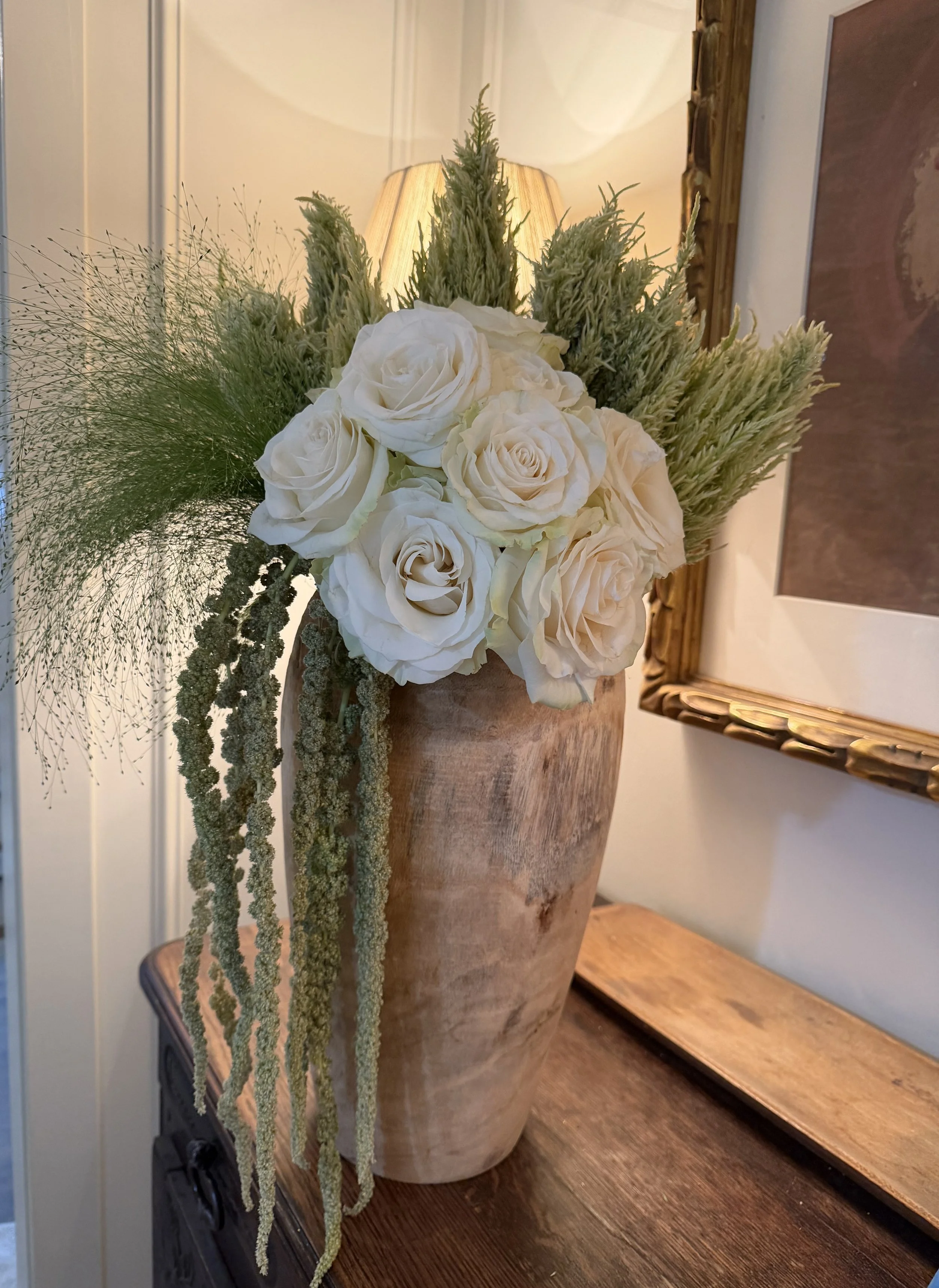 A tall beige vase holding a floral arrangement of white roses, green foliage, and hanging green strands, with a table lamp in the background and framed artwork on the wall.