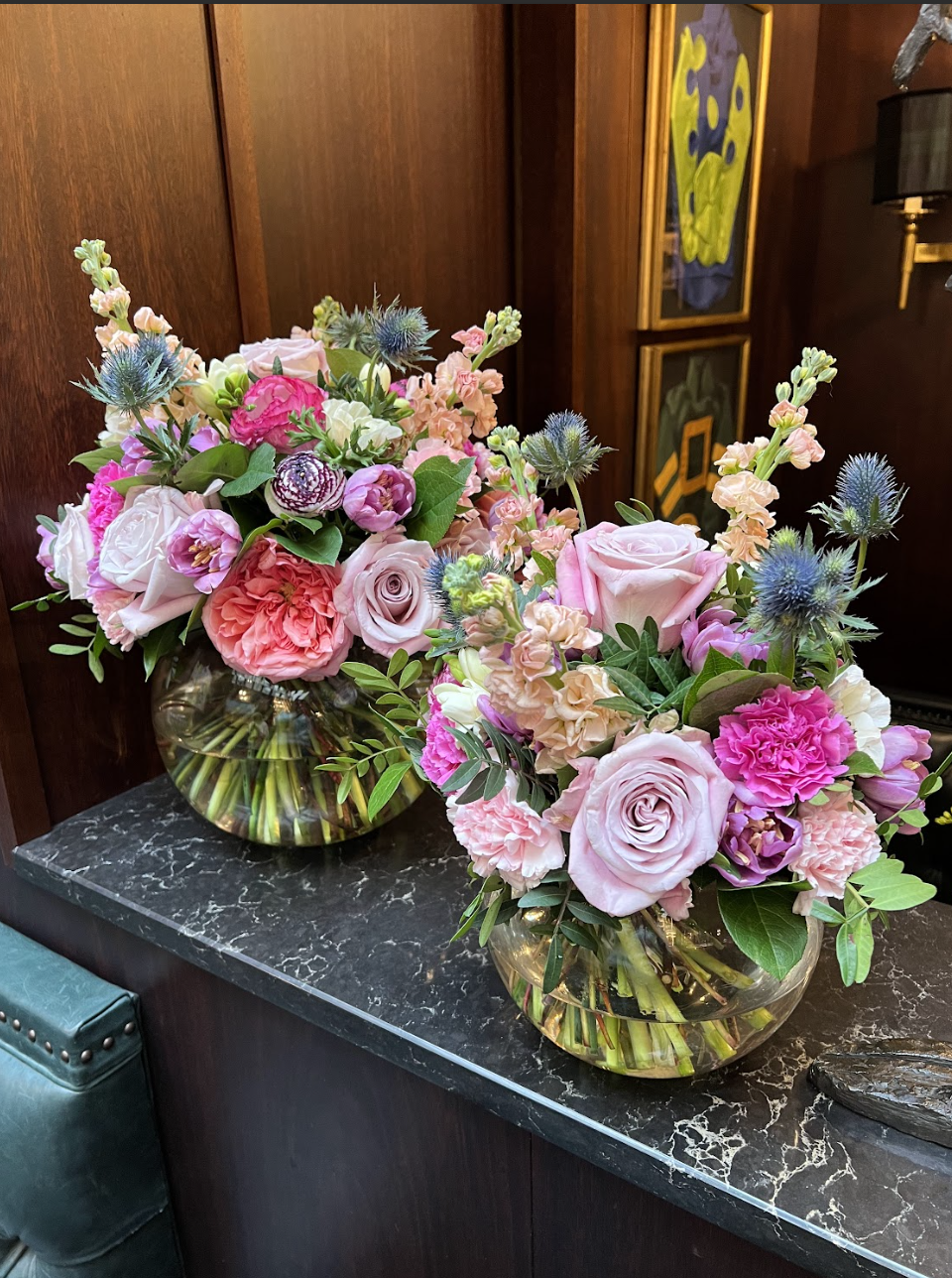 Two large glass vases filled with pink, white, and purple flowers, including roses, carnations, and thistles, placed on a black marble tabletop against a wooden wall.
