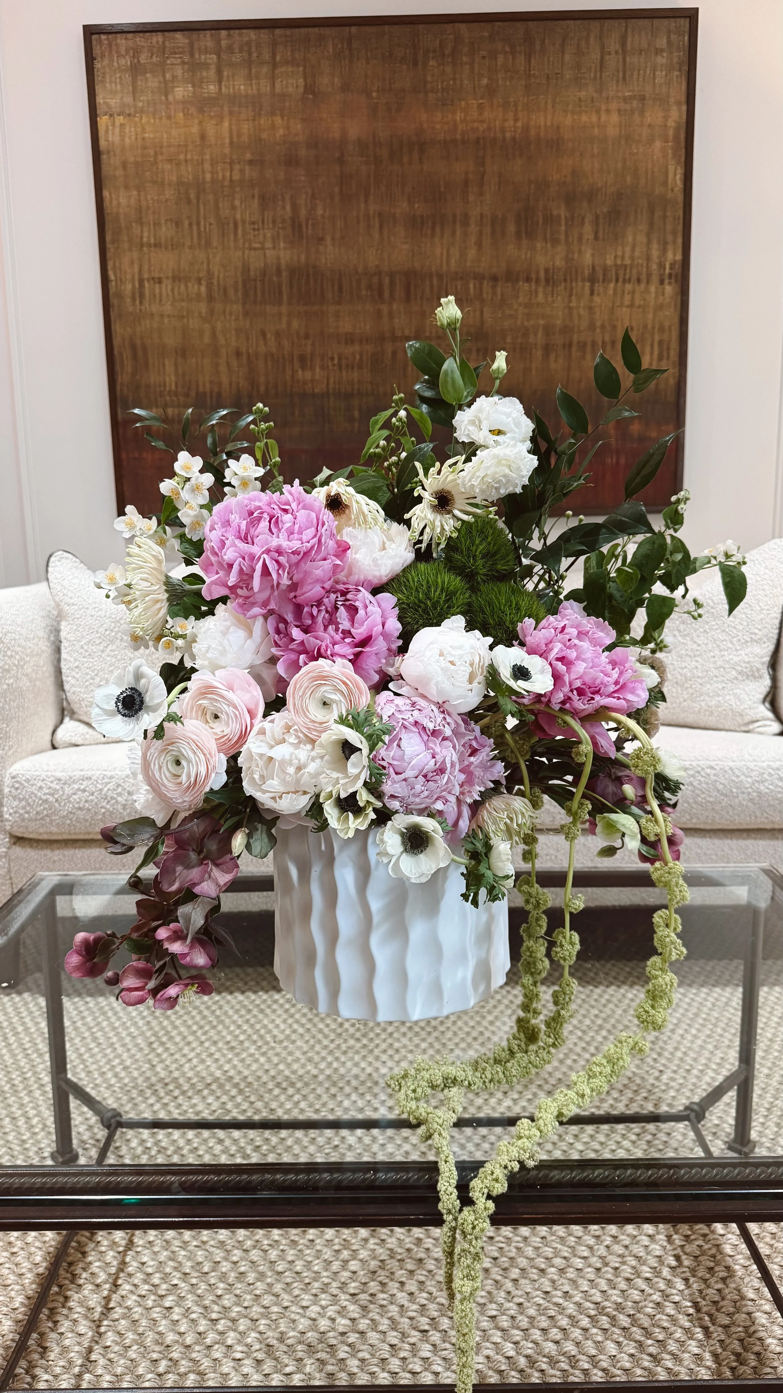 A large bouquet of pink and white flowers arranged in a white vase on a glass table in a room with beige carpeting, a beige sofa, and a wooden wall hanging.