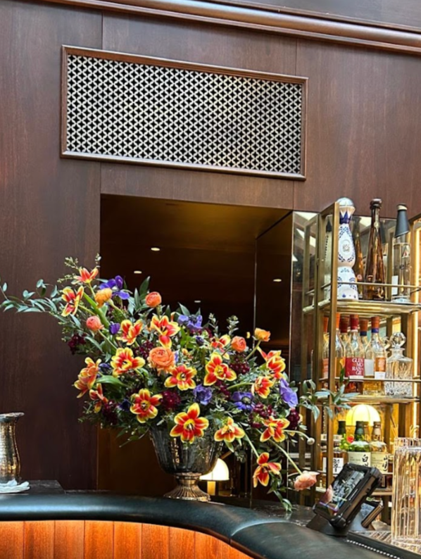 A floral arrangement with colorful flowers on a bar counter next to a liquor shelf in a dimly lit bar or restaurant.