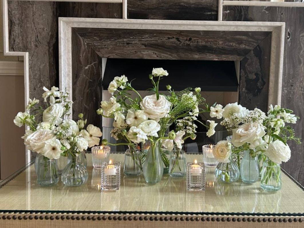 Multiple glass vases with white flowers on a glass-topped table, with candles in glass holders, in front of a fireplace.