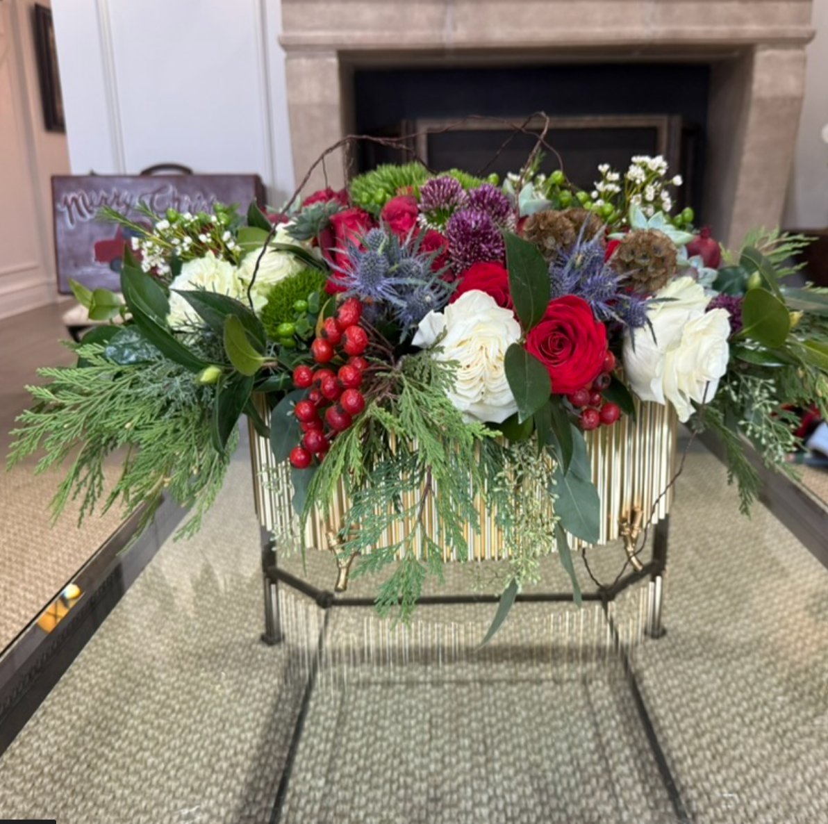 A festive floral arrangement with red, white, and purple flowers, green foliage, and red berries in a gold and black decorated container, placed on a table in a living room with a fireplace in the background.