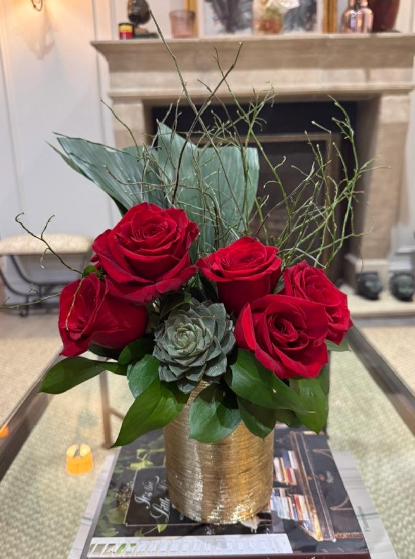 A floral arrangement in a gold vase with red roses, a succulent, large green leaves, and curly twigs, placed on a table in a cozy living room.