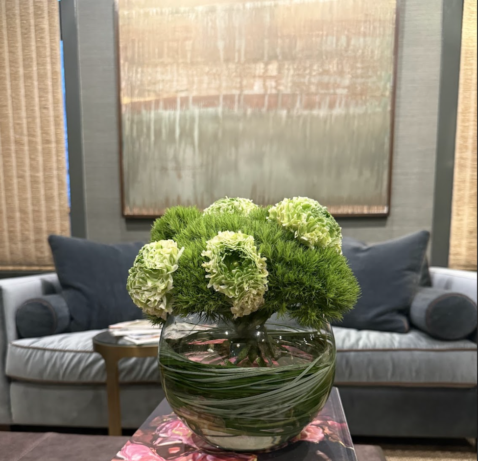 A glass vase with a floral arrangement of green and white flowers on a table in a living room with a sofa and a wall hanging in the background.