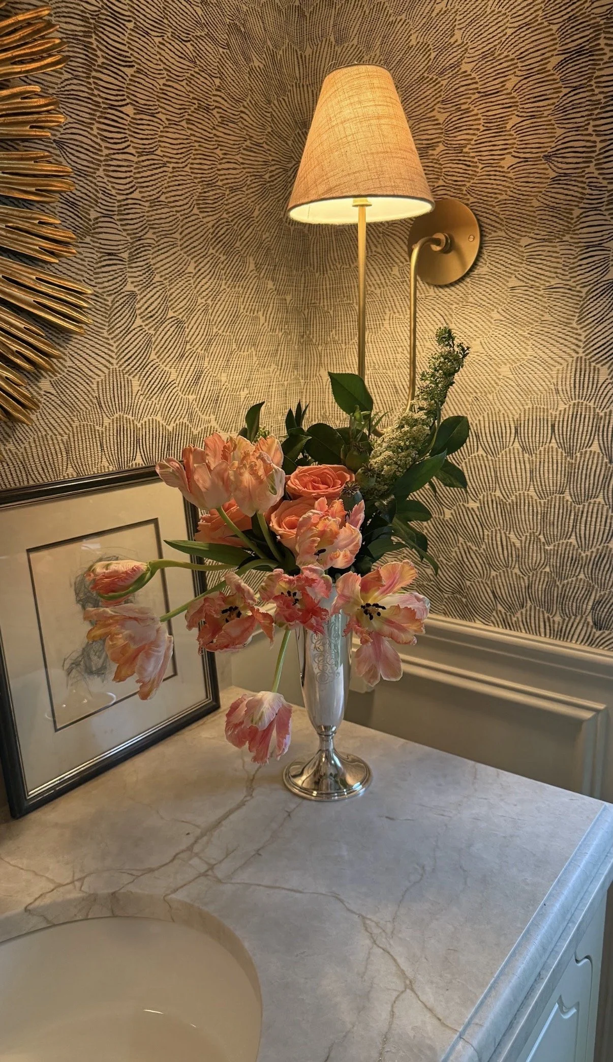 A bathroom countertop with a marble surface, a silver vase holding pink and peach flowers, a framed artwork, a wall-mounted lamp with a beige shade, a patterned wallpaper background, and part of a sink is visible.