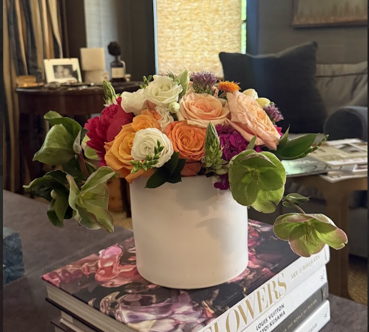 A bouquet of colorful flowers in a white vase, placed on top of several large books on a table in a cozy living room.