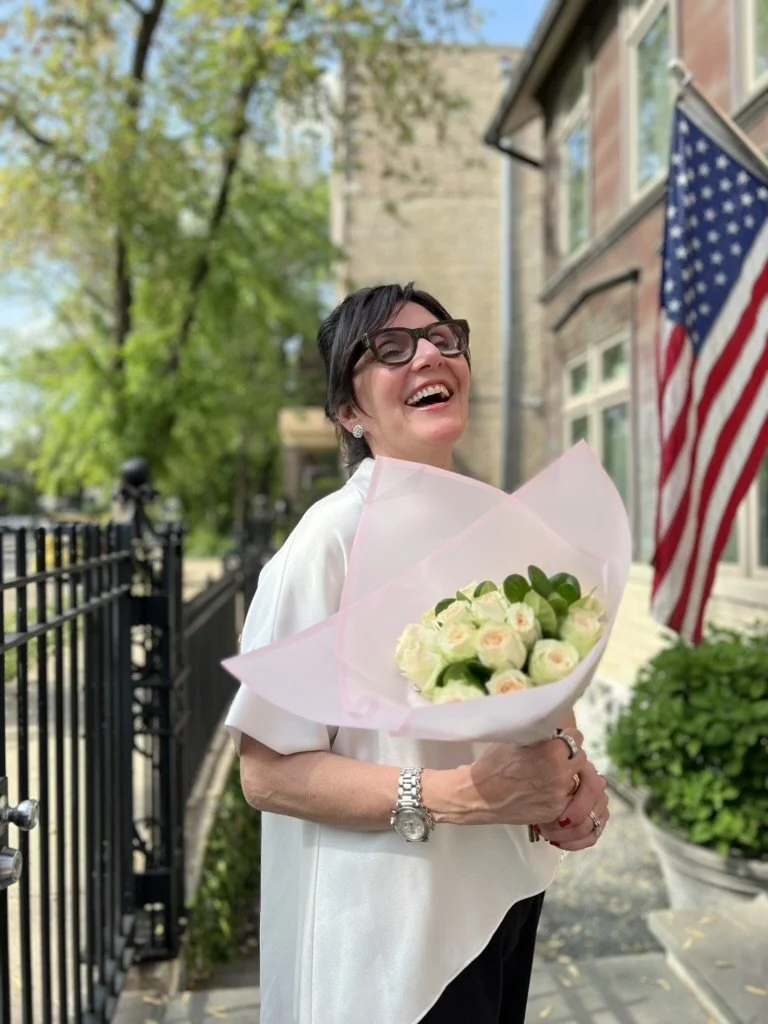 A woman smiling and holding a bouquet of white roses wrapped in pink paper, standing outside in front of a house with an American flag, with green trees and a black fence in the background.