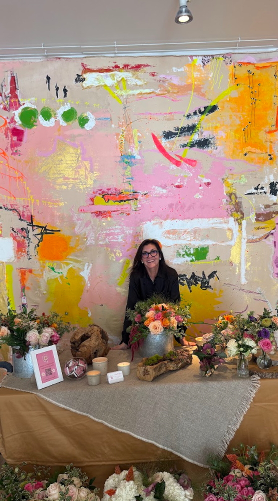 A woman sitting behind a flower display at a decorated table, with a colorful abstract mural on the wall behind her.