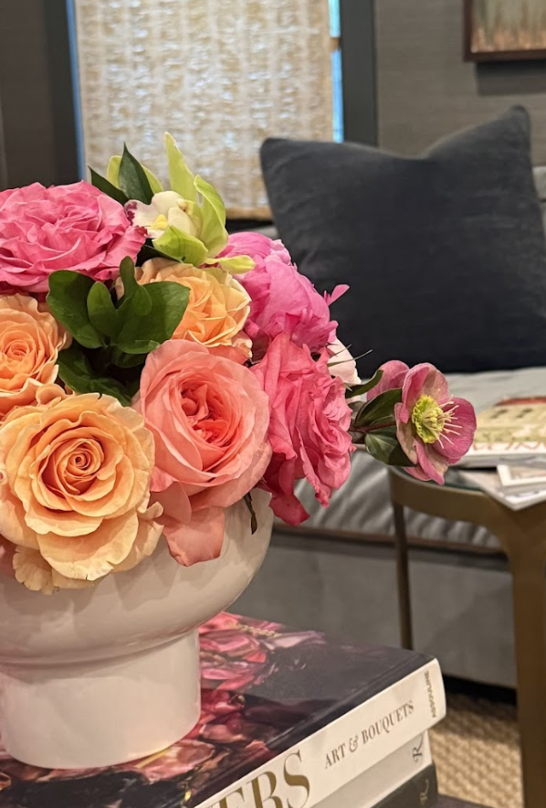 A white vase filled with pink, peach, and white roses, with some green leaves, placed on a book titled 'FLORAL ART & BOUTIQUE' in a cozy interior setting.