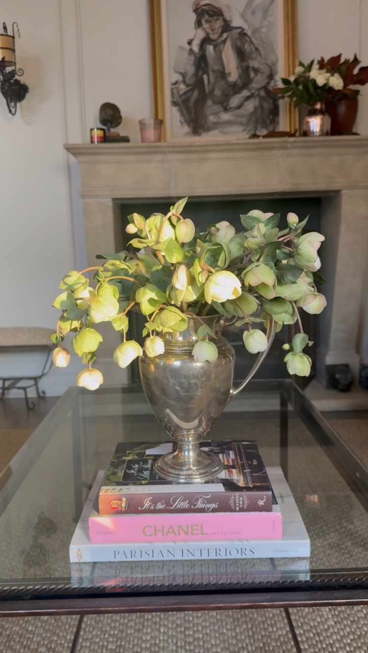 A glass vase with a bouquet of green and pinkish flowers is placed on top of three stacked books on a glass table. The books are titled 'Parisian Interiors,' 'Chanel,' and 'As the Little Things.' In the background, there is a fireplace with a framed 