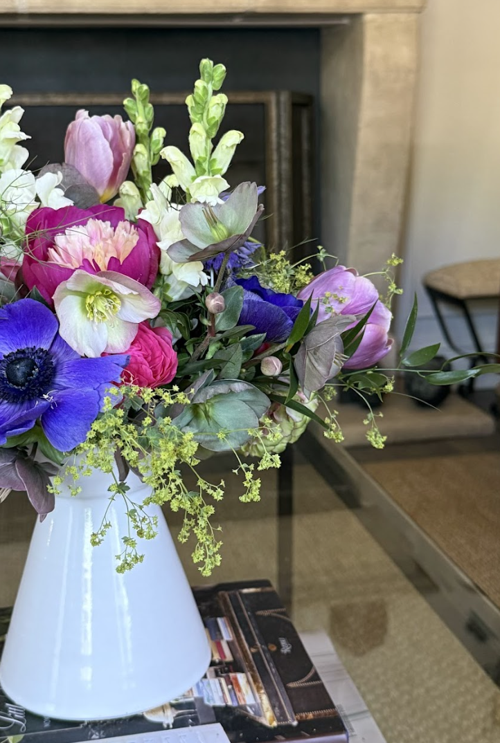 A colorful bouquet of flowers including pink, purple, and white blooms, arranged in a white vase on a glass table in a room with a fireplace and a chair in the background.