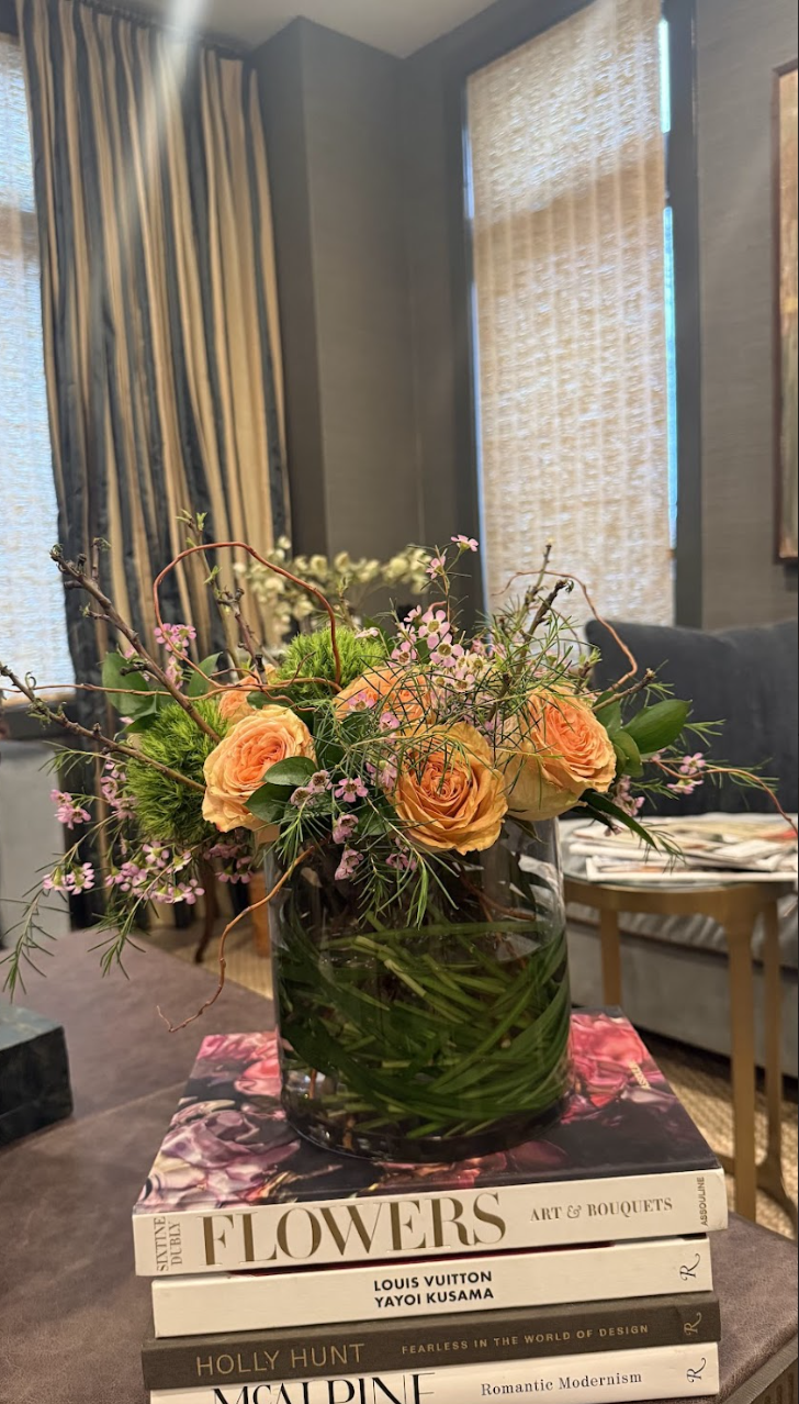 A floral arrangement with peach roses, small pink flowers, green foliage, and twigs in a clear glass vase placed on top of a stack of art books inside a room with curtains and a sofa.