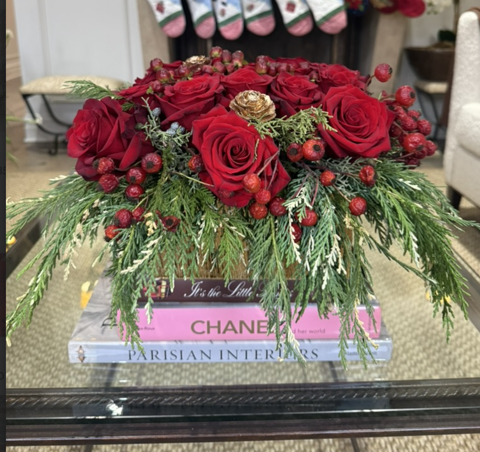 A floral arrangement with red roses, red berries, pine, and greenery, placed on top of a stack of fashion books including Chanel and Parisian Interiors.