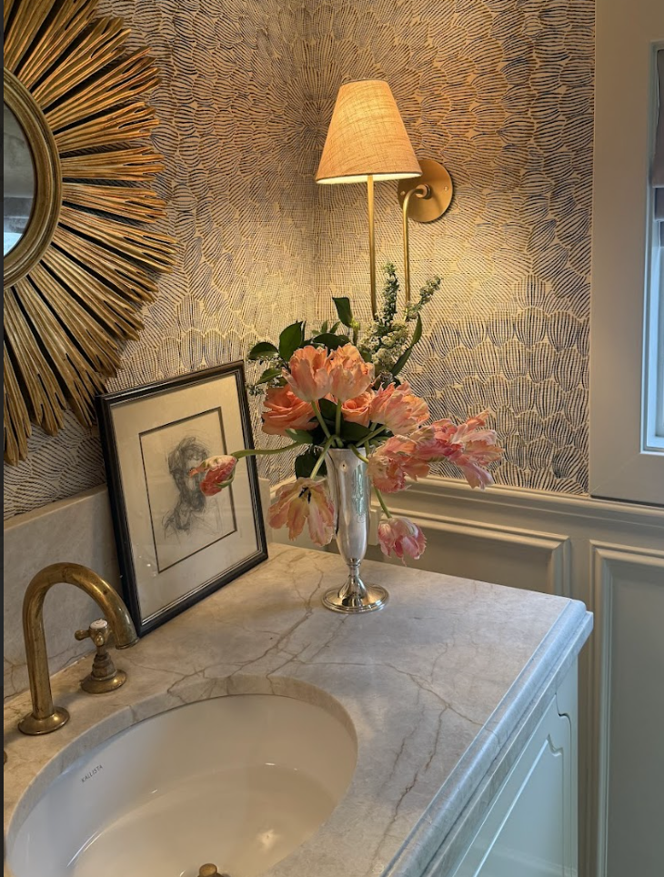Decorative bathroom corner with a marble countertop, a framed sketch leaning against a patterned wallpaper, a vase with pink flowers, a brass faucet, a wall-mounted lamp with a beige shade, and part of a mirror with a sunburst frame.