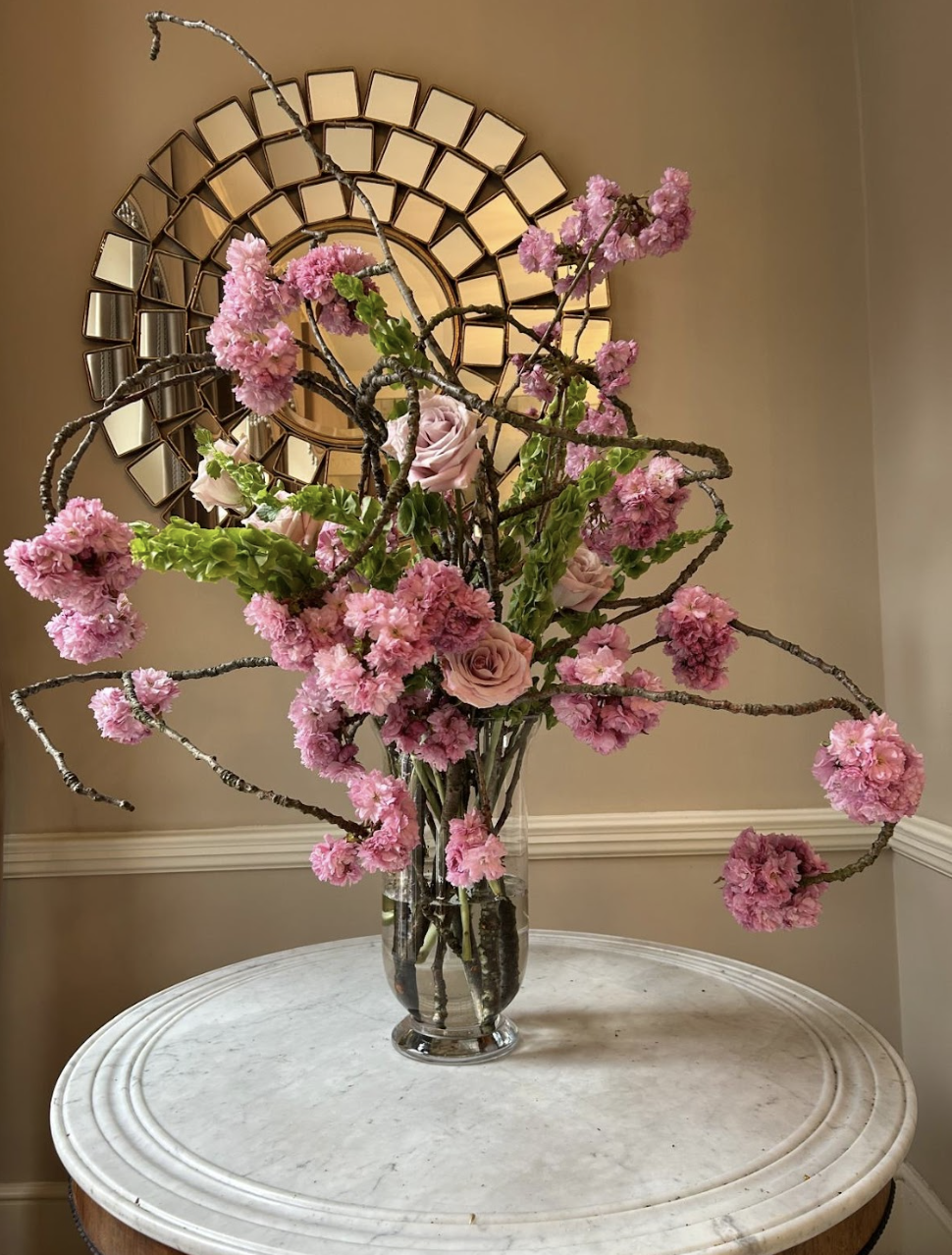 A tall glass vase filled with pink cherry blossoms and light pink roses, placed on a white marble table with a decorative gold mirror on the wall behind.
