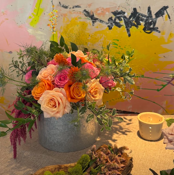 A floral arrangement with pink, orange, peach, and purple roses in a metallic gray vase, with green foliage and decorative plants. There is a small yellow candle holder and some natural elements on a beige table, with a colorful abstract wall in the 