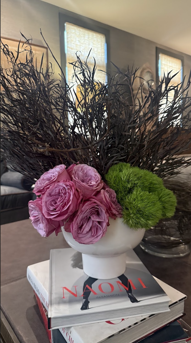 A white vase containing pink roses, green moss, and dark brown branches, placed on top of a fashion magazine on a table in a living room.