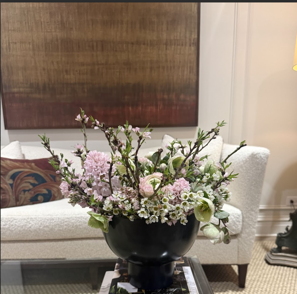 A black vase with pastel pink and white flowers, including lilies, arranged in a floral centerpiece on a glass table in a living room.