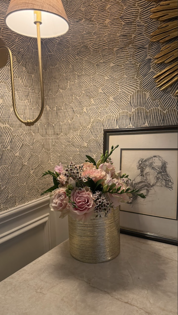 A vase with pink and white flowers on a beige tabletop, next to a framed black-and-white line sketch of a woman with curly hair, against patterned wallpaper, with a wall sconce and a gold decorative piece hanging on the wall.