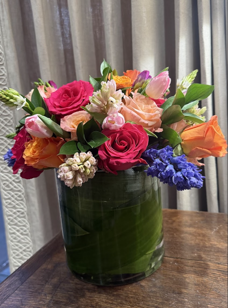 A colorful bouquet of flowers in a glass vase, placed on a wooden surface with curtains in the background.