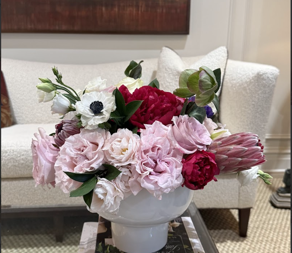 A bouquet of pink, white, and red flowers in a white vase on a table in front of a beige sofa.