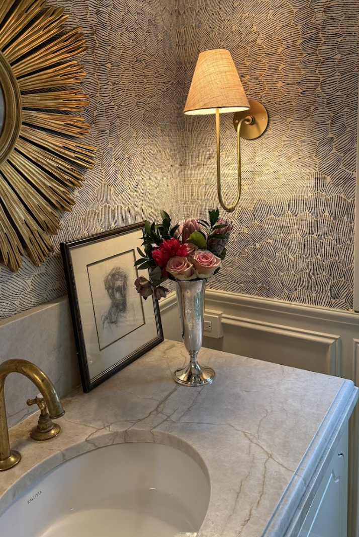 A bathroom sink vanity with a marble countertop. On the countertop are a framed sketch of a person, a silver vase with pink and red flowers, and a wall-mounted light fixture with a beige shade. The wall features textured wallpaper with a fan-shaped p