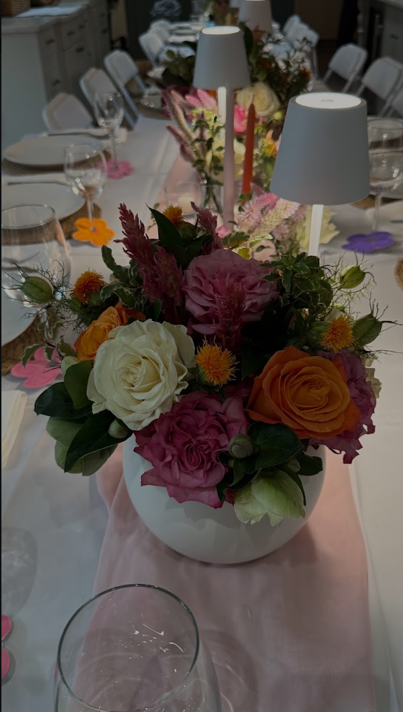 A floral centerpiece on a dining table featuring pink, orange, cream, and purple flowers in a white vase with two small white lamps and a pink table runner.