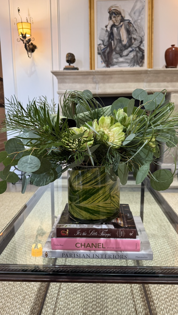 A floral arrangement in a green marbled vase sitting on top of three stacked books on a glass table in a stylish living room. The background features a fireplace, a painting of a person wearing glasses and a hat, a small sculpture, and a couple of va