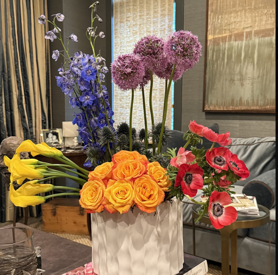 A bouquet of colorful flowers in a white textured vase, including yellow calla lilies, orange roses, purple alliums, and pink poppies in a cozy living room.
