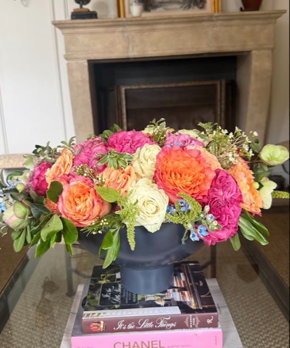 A colorful bouquet of pink, orange, cream, and purple flowers in a dark vase, placed on top of a stack of hardcover books in a living room with a fireplace in the background.