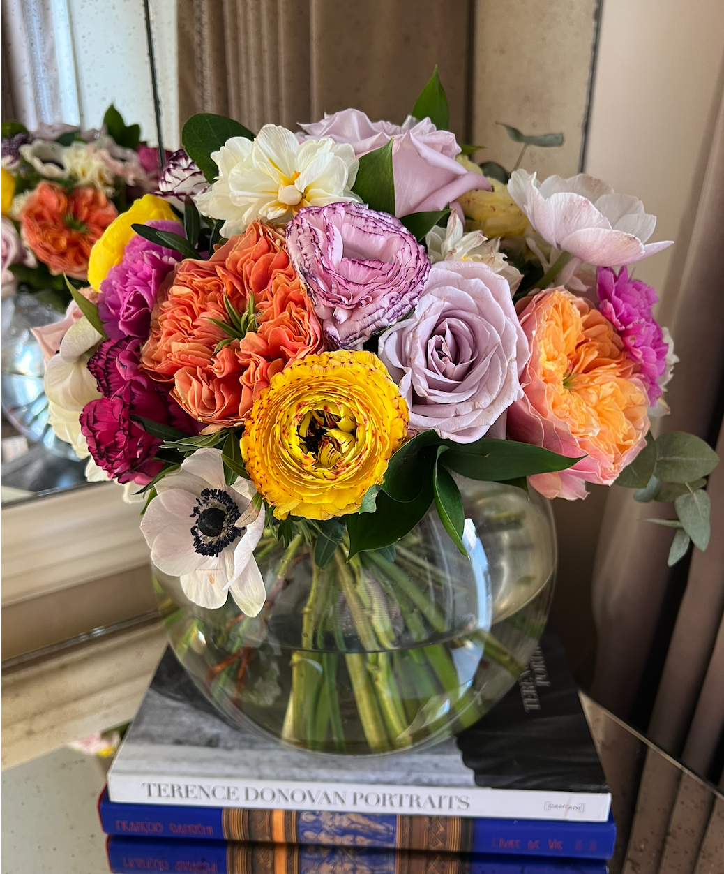 Colorful bouquet of various flowers in a round glass vase sitting on top of books on a table, near a mirror and curtains.