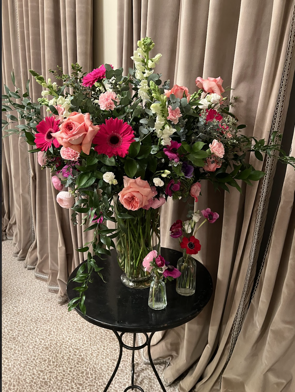 A large floral arrangement with pink, peach, white, and purple flowers in a glass vase, placed on a round black table against beige curtains.
