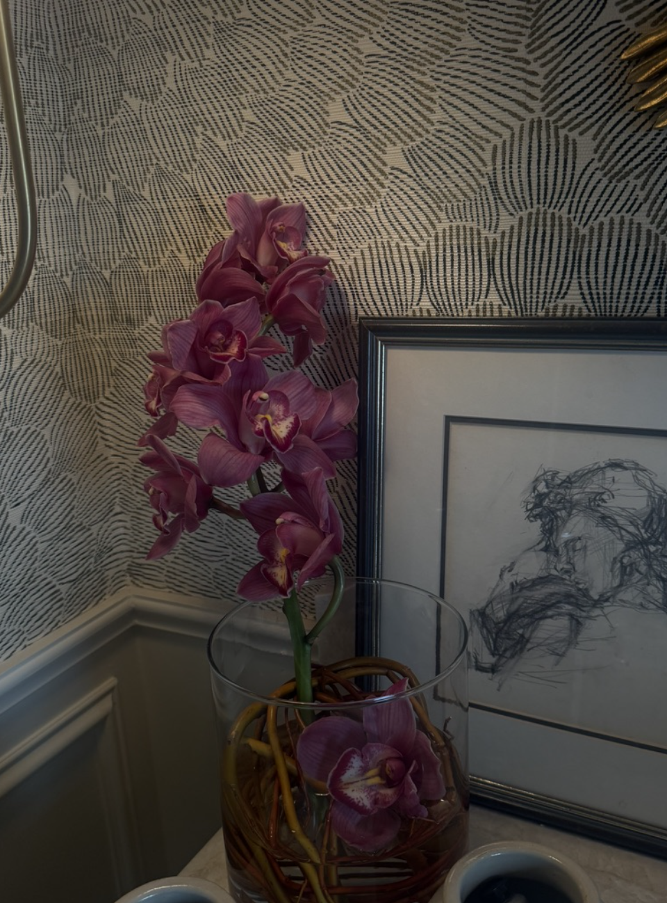 Pink orchid flowers in a glass vase next to a framed sketch of a dog, with patterned wallpaper in the background.