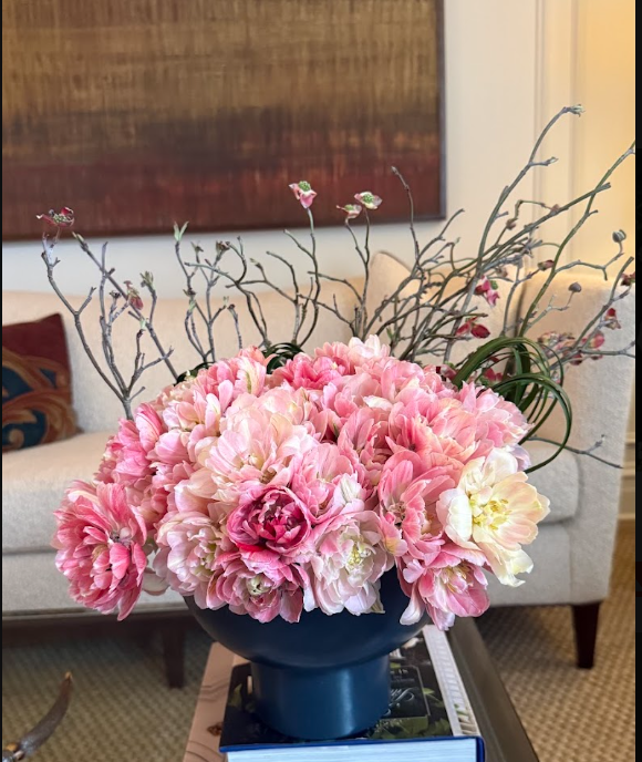 A floral arrangement of pink and cream flowers, including peonies and roses, with decorative twigs in a black vase on a coffee table in a living room.