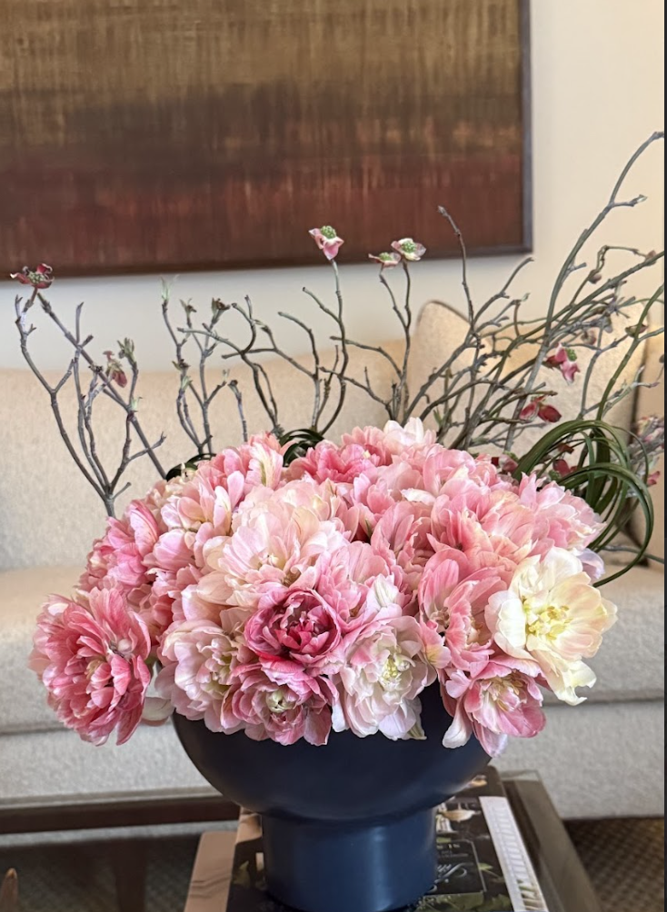 A black vase filled with pink and white peonies, some branches with small pink flowers, set on a table in a living room.