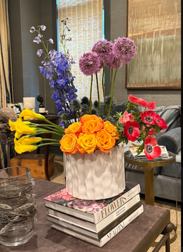 A bouquet of orange roses, yellow calla lilies, and pink poppies in a textured white vase, placed on top of a stack of four books about flowers and gardening, in a living room setting.