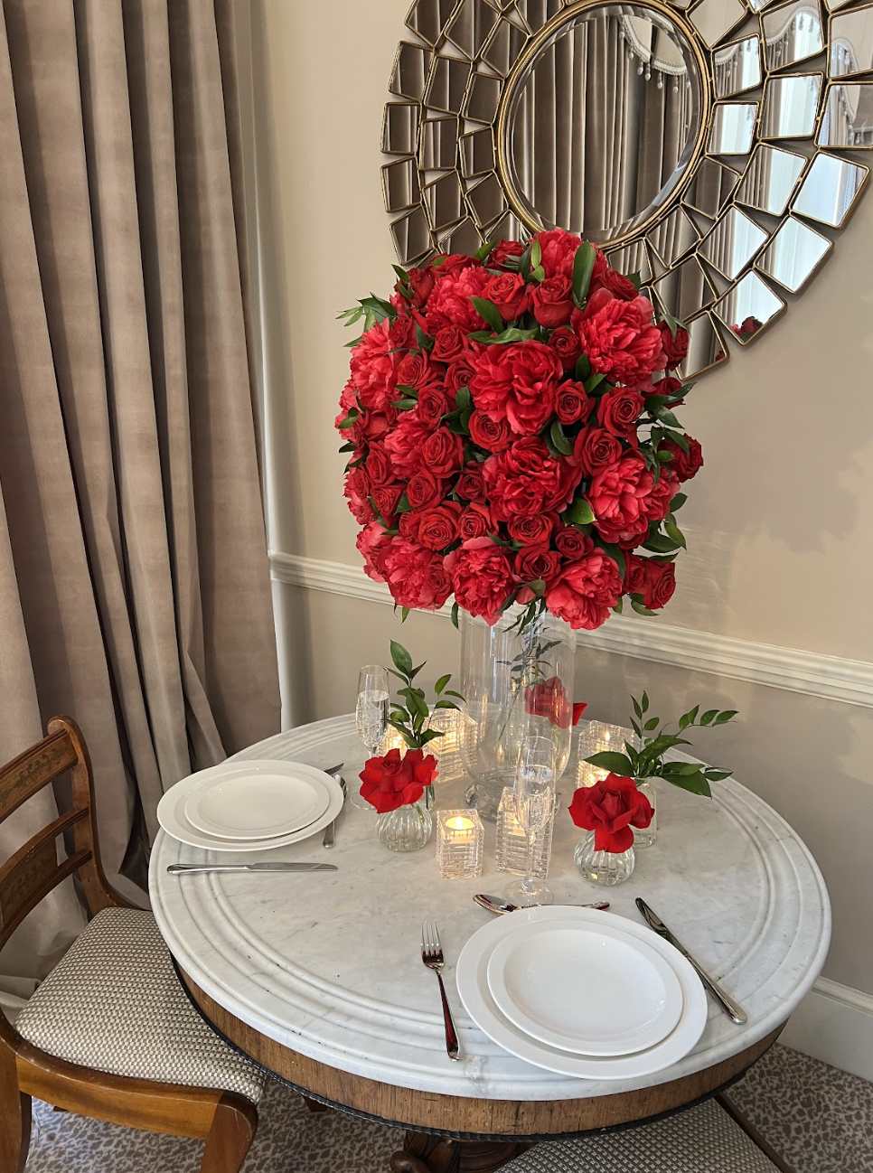 A decorated table with a large floral centerpiece of red roses and green leaves, surrounded by plates, glasses, and candles, in a dining room with beige curtains and a decorative mirror on the wall.