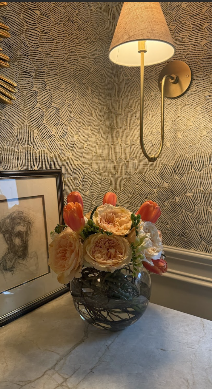 A floral arrangement in a glass vase with pink and orange flowers on a marble surface next to a framed sketch of a face and a wall-mounted lamp with a beige lampshade against a textured wallpaper.