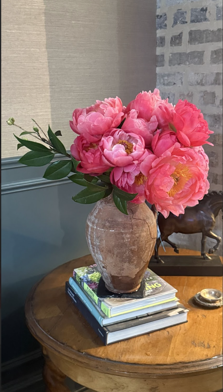A rustic clay vase filled with bright pink peony flowers and green leaves, placed on a stack of books on a round wooden table in a room with a brick wall and a textured wall.