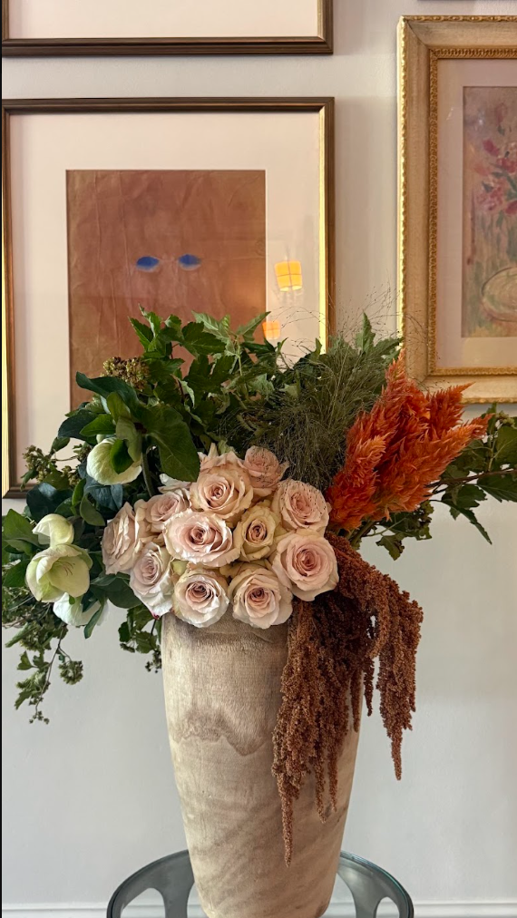 A tall beige vase holding a mixed floral arrangement, including pale pink roses, white hellebores, green foliage, and orange dried flowers, set against a wall with several framed artworks.