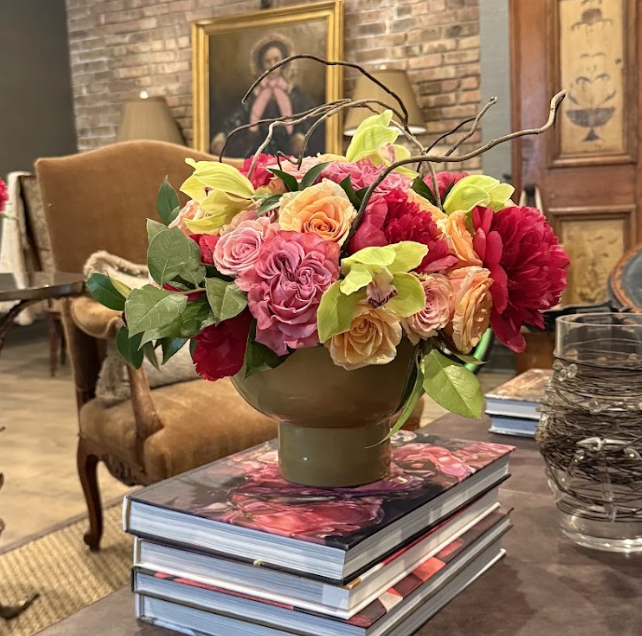A floral arrangement of pink, orange, yellow, and red flowers in a green vase on top of a stack of books on a wooden table.