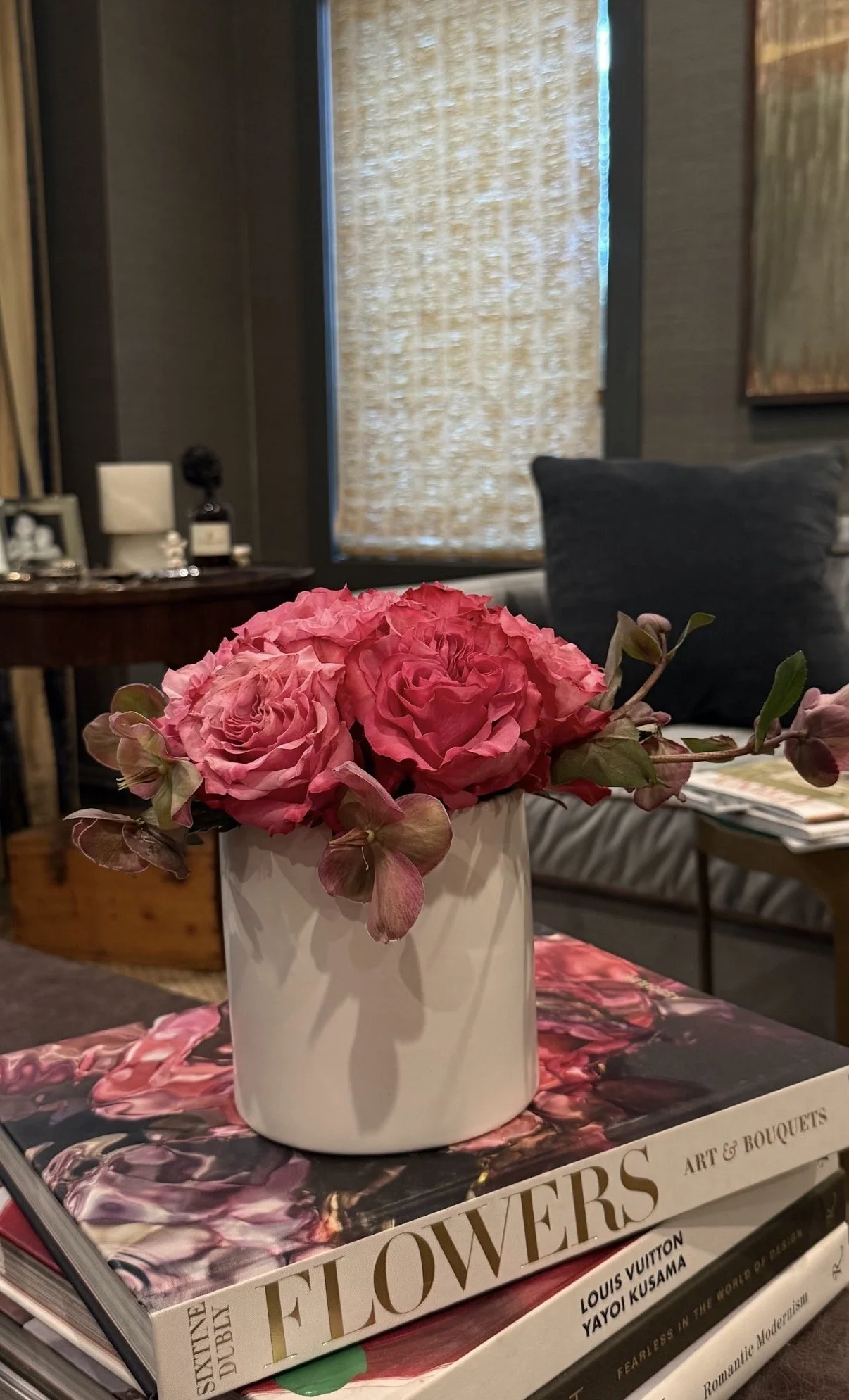 A white vase with pink flowers and green leaves on top of a book titled "Flowers Art & Bouquets" in a cozy living room with dark walls, a window with bamboo curtains, and a dark sofa.
