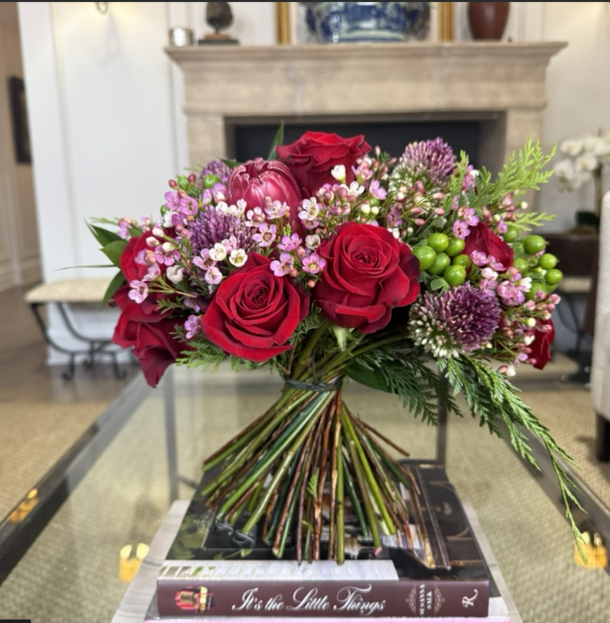 A bouquet of red roses, pink wax flowers, purple alliums, green hypericum berries, and ferns in a glass vase on a table.