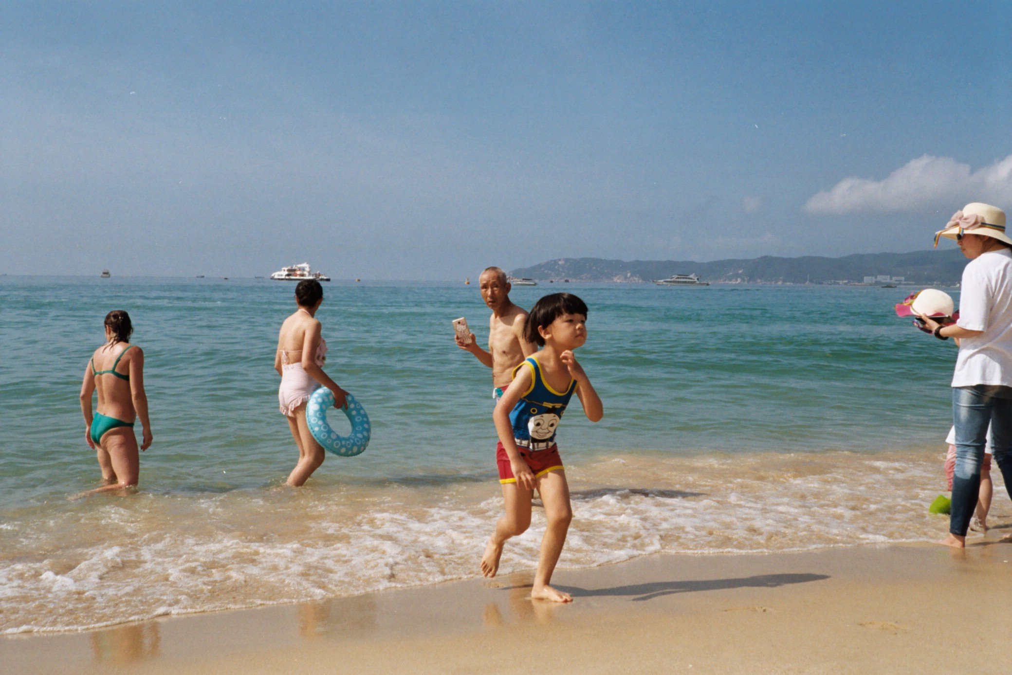 People enjoying the beach near the water shoreline, with children and adults in swimwear and on the sand, and boats in the distance under a partly cloudy sky.