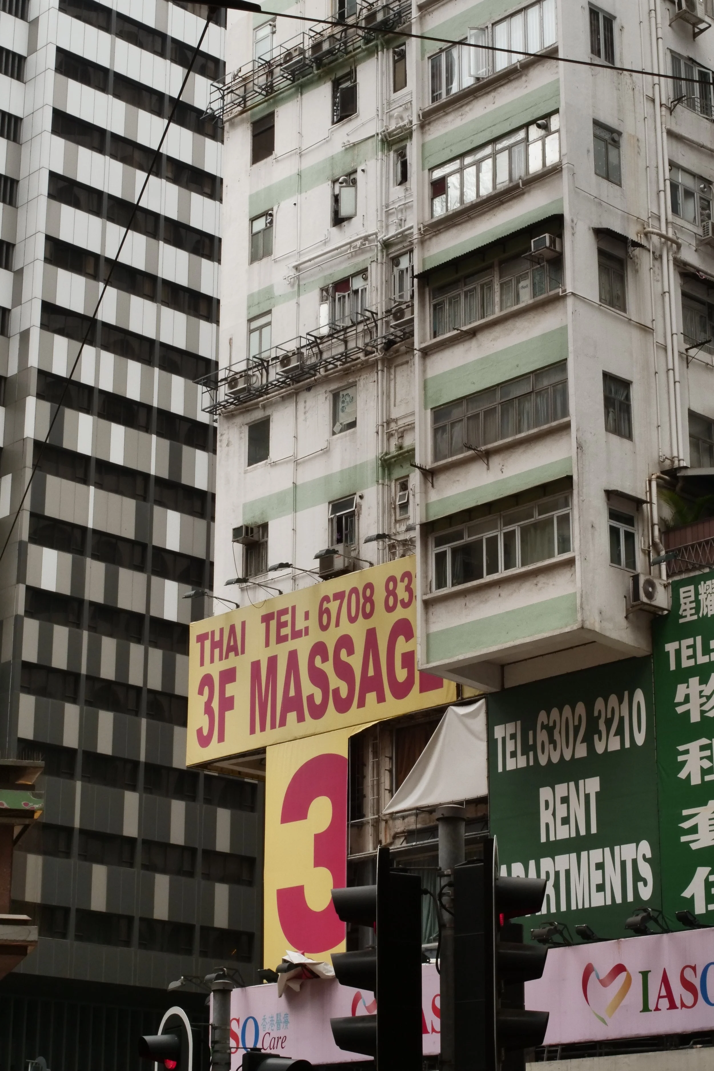 A tall building with multiple windows and air conditioning units. A large yellow sign with pink lettering advertises a Thai massage on the third floor, with a phone number. Other signs below advertise rent apartments and other services.