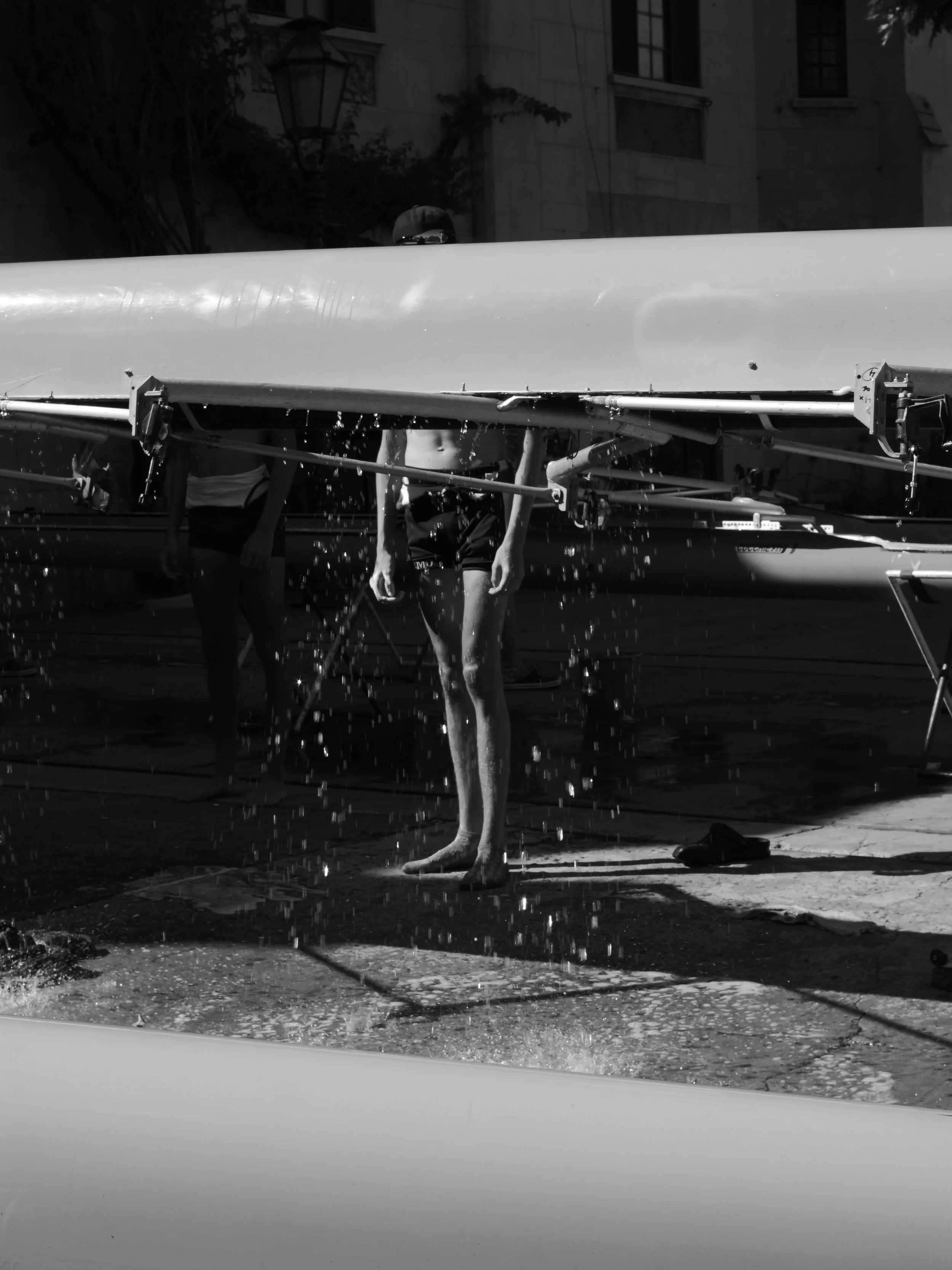 Two rowers in sportswear cleaning or preparing their boat, with water splashing on the pavement in an urban setting.