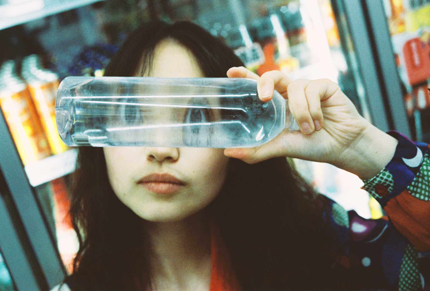 A woman with dark hair standing in front of vending machines, holding a clear plastic water bottle up to her eye as if to look through it.