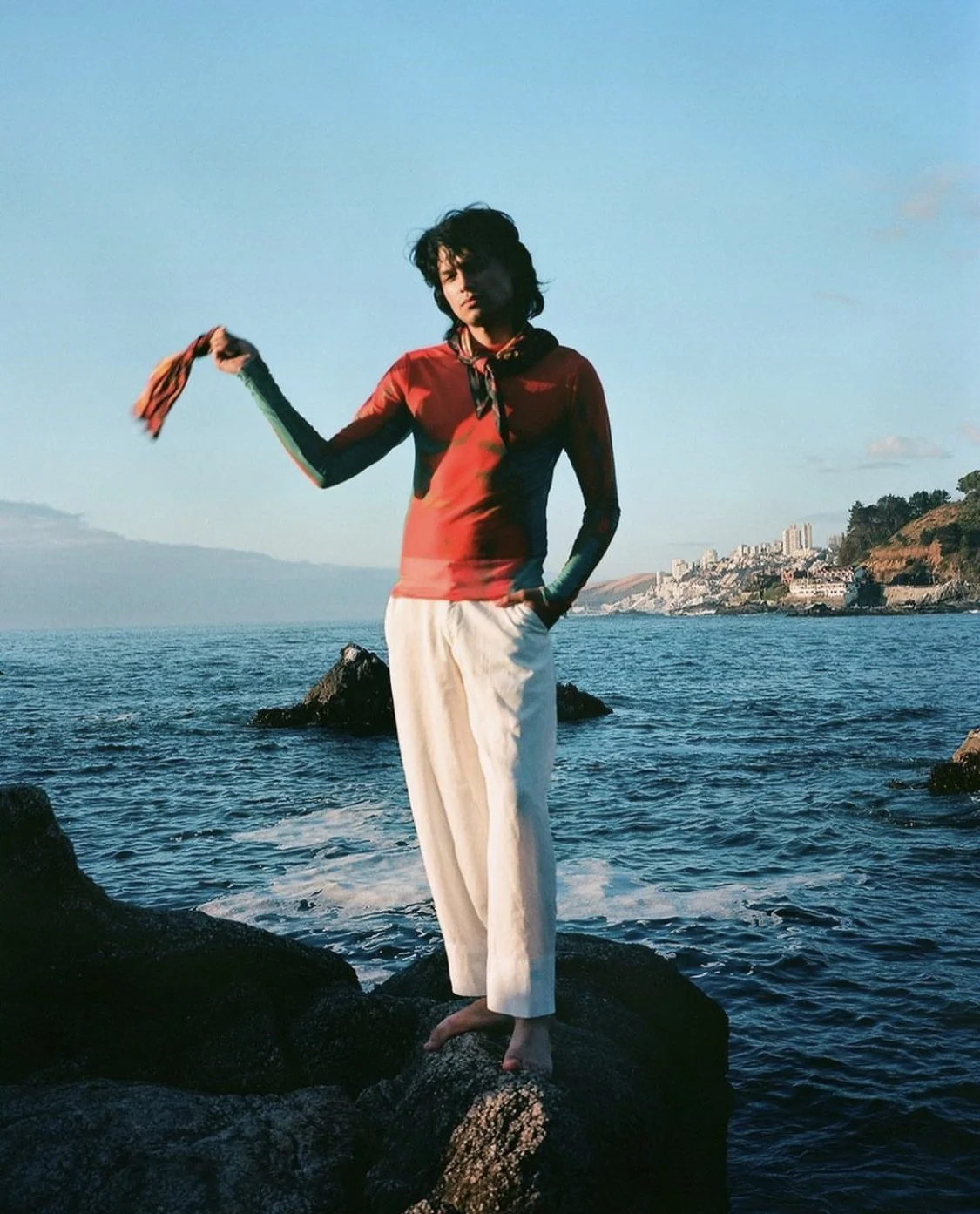 A woman standing barefoot on rocks in the ocean, holding a cloth in her right hand, with a cityscape and hills in the background under a clear sky.