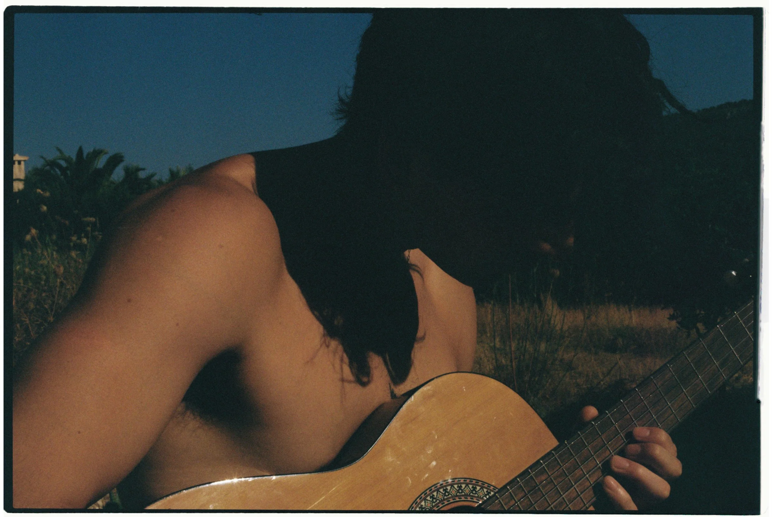 A person with long dark hair, wearing a sleeveless top, playing an acoustic guitar outdoors during dusk or nighttime with a landscape of trees and sky in the background.